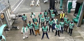 Eine Gruppe von etwa 23 Personen steht im Freien, tragen grüne Westen mit Logo. Industrielle Ausrüstung im Hintergrund.