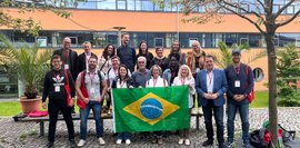 Vor dem Hochschulgebäude lächeln ca. 20 Personen mit brasilianischer Flagge. Vielfältige Gruppe, informelle Kleidung.