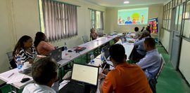 Hier findet ein Planungsworkshop statt. Eine Gruppe von Menschen sitzt an einem langen Tisch und schaut auf eine Leinwand mit einer Präsentation. Es sind Laptops und Wasserflaschen zu sehen. Der Raum wirkt hell und freundlich.