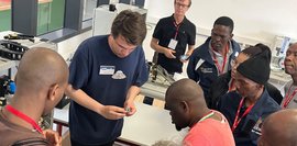 Die Teilnehmer der Delegationsreise schauen sich die Austattung in einer Ausbildungswerkstatt an. Eine Gruppe von Menschen steht im Kreis um einen Tisch auf dem verschiedene Materialien der Ausbildungswerkstatt liegen. Eine Person erklärt ein mechanisches Teil, welches er in der Hand hält.