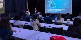 Konferenzraum: Vortrag über grüne Wasserstofftechnologie vor Publikum. Präsentation mit H2-Logo auf Leinwand.