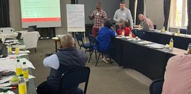 Workshop von Unternehmen und Colleges Einige Menschen sitzen und stehen in einem Seminarraum. Eine Person präsentiert am Flipchart.