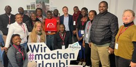 Gruppe von ca. 15 Personen, hauptsächlich Frauen und Männer afrikanischer Herkunft, posieren vor einem Banner mit der Aufschrift „Wir machen Urlaub! Tourismusbranche Bayern“. Ein Mann im Anzug steht im Zentrum. Freundliche, positive Atmosphäre.