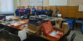 In einem Schulungsraum stehen mehrere Personen um einen Tisch mit Werkzeugkisten, Laborgeräten und technischer Ausrüstung. Einige Teilnehmer zeigen mit dem Daumen nach oben.