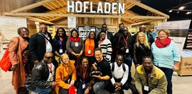 Große Gruppe lächelnder Menschen vor einem Hofladen-Stand. Unterschiedliche Altersgruppen und Ethnien sind vertreten. Freundliche, einladende Atmosphäre.