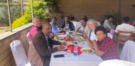 Hier genießen mehrere Personen ein gemeinsames Essen im Restaurant des Selam Colleges. Angeordnet an Tisch und Stühlen, sind sie in angeregter Stimmung. Sonnenlicht fällt auf die Szene.