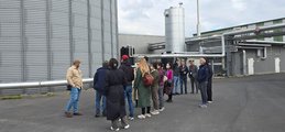 Eine Gruppe Leute steht vor einem großen Silo und anderen Anlagen einer Biogasanlage. Ein Mann führt die Gruppe.