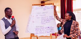 Eine Frau hält ein Plakat hoch, während ein Mann mit Mikrofon die Inhalte erläutert.