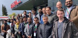 Vor dem RAPA-Gebäude steht eine große Gruppe von Menschen, hauptsächlich Männer, in formeller Kleidung. Sie lächeln in die Kamera. Der Himmel ist bewölkt. Ein freundliches Gruppenfoto.