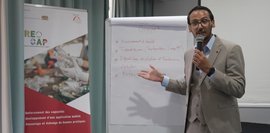 Eine Person steht vor einem Flipchart und erklärt Inhalte. Links ist ein Roll-up mit dem RECAP-Logo und Projektinformationen sichtbar.