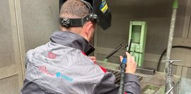Ein Mann mit einem hochgeklappten Schweißhelm sitzt auf einem Hocker in einer Metallkabine und hält ein Schweißgerät in der Hand.