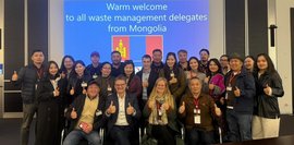 Eine Gruppe mongolischer Abfallmanagement-Delegierter posiert lächelnd mit erhobenem Daumen vor einer Leinwand mit der Aufschrift "Warm welcome".