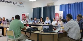 Besprechung: Menschen sitzen an einem Tisch in einem Konferenzraum, trinken und diskutieren. Laptop sichtbar.