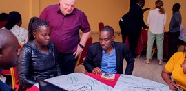 Mehrere Personen sitzen um einen Tisch und besprechen Inhalte auf einem großen Plakat, während ein Mann im roten Hemd zuhört.