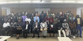 Eine große Gruppe von Menschen posiert für ein Foto in einem hell erleuchteten Raum. Die meisten sitzen oder stehen.