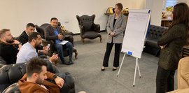 Bild zeigt einen Workshop zum Projektmanagement in Erbil. Eine Frau präsentiert vor einer Gruppe von Männern, die auf Ledersofas sitzen. Ein Flipchart steht im Raum. Der Raum ist modern eingerichtet.