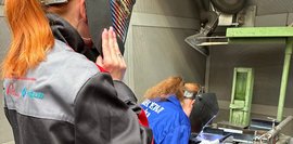 Eine Frau mit hochgeklapptem Schweißerhelm beobachtet eine andere Person, die in blauer Arbeitskleidung ein Metallstück schweißt. Die Szene findet in einer Werkstattkabine mit Belüftungssystem statt.