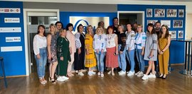 Eine Gruppe von Frauen steht in einem modernen Gebäude mit blau-weißer Wandgestaltung und Unternehmenslogos von AZ Zwickau. Sie tragen traditionelle ukrainische Blusen und moderne Kleidung und blicken in die Kamera.
