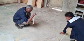 Zwei Männer messen mit einem Maßband auf einem Betonboden in einer Werkstatt aus. Der Boden ist abgenutzt und zeigt Farbflecken. Holz liegt herum. Die Männer hocken, konzentriert bei der Arbeit.