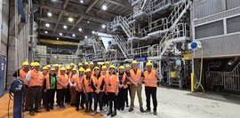 Eine Gruppe trägt Sicherheitshelme in einer Papierfabrik. Große Maschinen sind im Hintergrund. Der Boden ist orange. Alle lächeln in die Kamera. Ein informativer und freundlicher Gruppen-Ausflug.