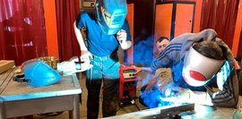 Zwei junge Männer mit Schweißhelmen arbeiten an einem Metallstück, während ein blauer Lichtschein vom Schweißen sichtbar ist.