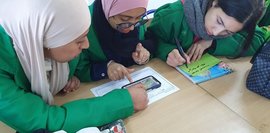 Drei junge Frauen in grünen Jacken sitzen an einem Tisch und arbeiten gemeinsam an einem Projekt. Sie betrachten ein Smartphone, schreiben Notizen und scheinen sich auszutauschen. Zusammenarbeit und Lernen im Vordergrund.