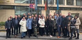Eine Gruppe von Männern und Frauen steht vor einem modernen Gebäude mit Glasfassade. Im Hintergrund sind die deutsche und die ukrainische Flagge gehisst. Die Teilnehmer tragen warme Winterkleidung und schauen in die Kamera.