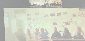 Bild zeigt einen Screenshot einer Videokonferenz. Eine Gruppe junger Menschen sitzt an einem Tisch vor einer Wand mit Bildern. Im Hintergrund weitere Teilnehmer. Der Bildschirm wirkt unscharf.