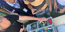 Draufsicht auf einen geöffneten Stromwechselrichter. Mehrere Personen betrachten die Elektronikkomponenten. Ein Mann erklärt das Innere.