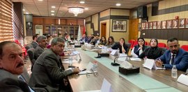 Gruppenfoto vom Feinplanungsworkshop 2024. Etwa 20 Personen sitzen an einem langen Konferenztisch in einem hellen Raum mit dunklem Mobiliar. An der Wand hängen Porträts und Fahnen.