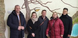 Fünf Personen in Winterkleidung posieren vor einer Wand mit einer dekorativen Baumstruktur. Sie lächeln in die Kamera.