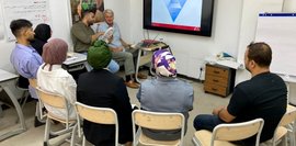 Hier findet ein Workshop mit Fachgruppen in Bagdad statt. Ein Mann präsentiert an einer Leinwand. Teilnehmer*innen sitzen in einem Raum und verfolgen aufmerksam die Präsentation. Diskussion und Austausch stehen im Mittelpunkt.