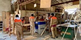 Hier arbeiten mehrere Männer in einer Holzwerkstatt an Holzarbeiten. Sie tragen beige Arbeitskleidung mit orangenen Details. Im Hintergrund befinden sich Maschinen und diverses Holz. Der Raum ist weitläufig und wirkt eher rustikal.