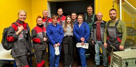 Eine Gruppe von Männern und Frauen, einige in blauen oder roten Schutzanzügen, andere in Alltagskleidung, steht in einer Werkstatt mit gelben Wänden und schaut in die Kamera.