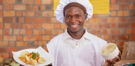 Lächelnder Koch in weißer Kochkleidung vor Ziegelwand hält Teller mit gebratenem Fisch und Gemüse sowie eine Schüssel mit einer grünen Soße. Er strahlt Stolz und Freude aus.