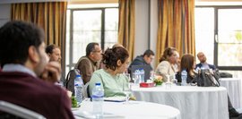 Konferenzraum mit mehreren runden Tischen. Es sitzen diverse Personen an den Tischen. Wasserflaschen und Gläser stehen bereit.