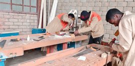 Drei junge Männer arbeiten konzentriert an Werktischen in einer Holzwerkstatt. Sie benutzen verschiedene Werkzeuge, um Holz zu bearbeiten. Der Raum ist einfach, aber funktionell eingerichtet. Ein Gefühl von Teamwork und handwerklicher Tätigkeit vermittelt das Bild.