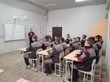 Ein Ausbilder spricht vor Auszubildenden in grauen und roten Uniformen in hellem Raum mit Whiteboard.