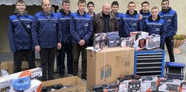 Eine Gruppe junger Männer in blauen Arbeitsanzügen steht vor einer Berufsschule, umgeben von neuen Werkzeugkoffern, Kartons mit technischer Ausrüstung und einem blauen Werkzeugschrank.