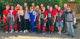 Eine Gruppe von Männern und Frauen steht vor einer grünen, mit Efeu bewachsenen Wand. Viele tragen rote und graue Arbeitskleidung mit Logos, andere sind in Freizeitkleidung. 