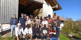 Vor einer Scheune posiert eine Gruppe Menschen. Es sind etwa 15 Personen unterschiedlichen Alters und Geschlechts, viele tragen rote Namensschilder. Heuballen sind im Hintergrund sichtbar. Sonniger Tag.