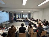 Teilnehmer sitzen an Tischen in einem hellen Raum. Zwei Personen referieren vor einer Leinwand.