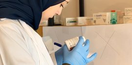 Apothekerin in weißem Kittel und dunklem Kopftuch prüft Medikamentenpackung. Sie trägt blaue Handschuhe und notiert etwas. Im Hintergrund sind Medikamentenschränke.