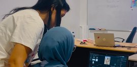 UTC-Personal mit Marketing-Trainerin Eine Marketing-Trainerin schaut einem Teammitglied des UTC über die Schulter, die vor einem Laptop sitzt.