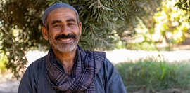 Lächelnder Mann mittleren Alters mit grauen Haaren und einem grauen Schal vor einem Olivenbaum. Er trägt eine graue Mütze. Das Bild strahlt Wärme und Ruhe aus.