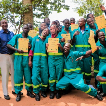 Freudig lächelnde Gruppe von Menschen in türkisfarbenen Arbeitsuniformen hält Zertifikate in Händen. Sie stehen draußen vor Bäumen. Erfolg und Ausbildung im Fokus.