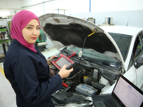 Eine weibliche Auszubildende mit Kopftuch steht vor einem Lehrauto und hält ein Diagnosegerät in den Händen.