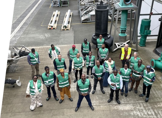 Eine Gruppe von Leuten trägt grüne Westen vor großen Rohren und Gerüsten.