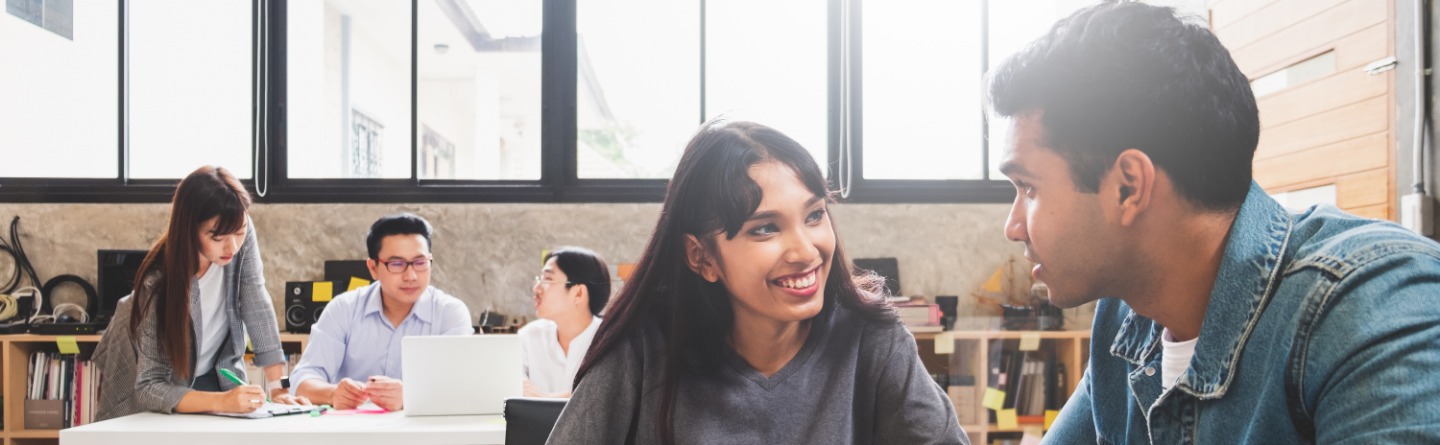 In einem lichtdurchfluteten Büro sitzen eine Frau und ein Mann an einem Tisch, sehen sich lächeld an. Vor ihnen ein offener Laptop. Im Hintergrund sind weitere Kollegen und eine große Fensterfront.