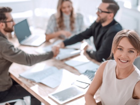 Geschäftspersonen sitzen bei einem Meeting. Im Hintergrund reichen sich zwei Männer die Hand. Eine Frau lächelnd einladend in die Kamera.